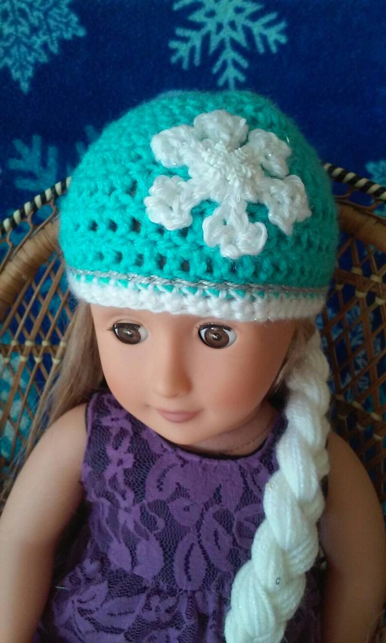 May include: A doll wearing a teal blue crocheted hat with a white snowflake design and a white crocheted braid.