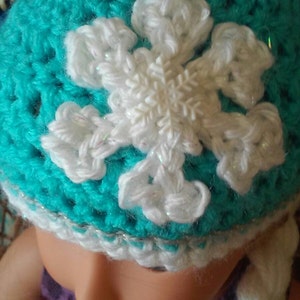 May include: A turquoise crocheted hat with a white crocheted snowflake design. The hat has a white crocheted braid attached to it.