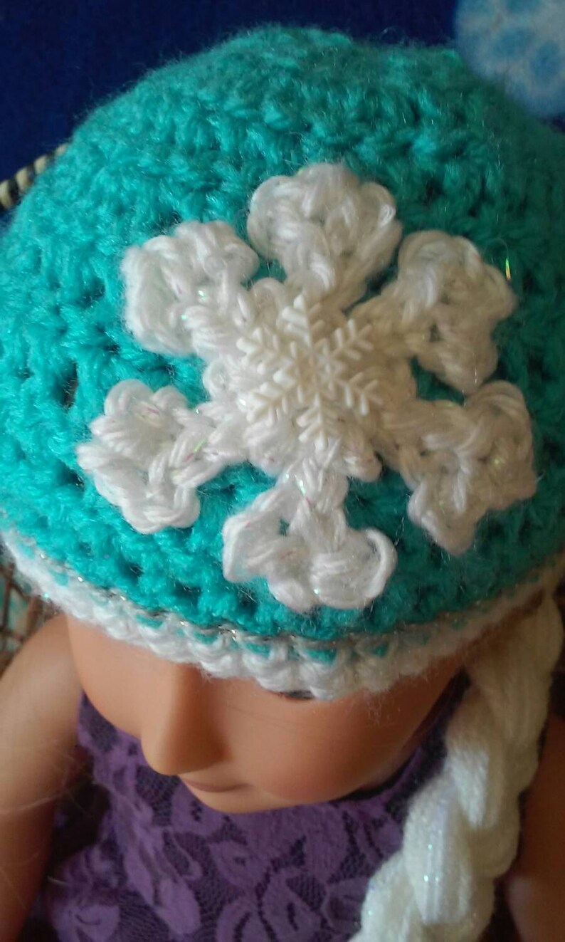 May include: A turquoise crocheted hat with a white crocheted snowflake design. The hat has a white crocheted braid attached to it.