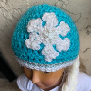 May include: A teal blue crocheted hat with a white snowflake design and a silver glitter trim. The hat is worn by a doll with long white braided hair.