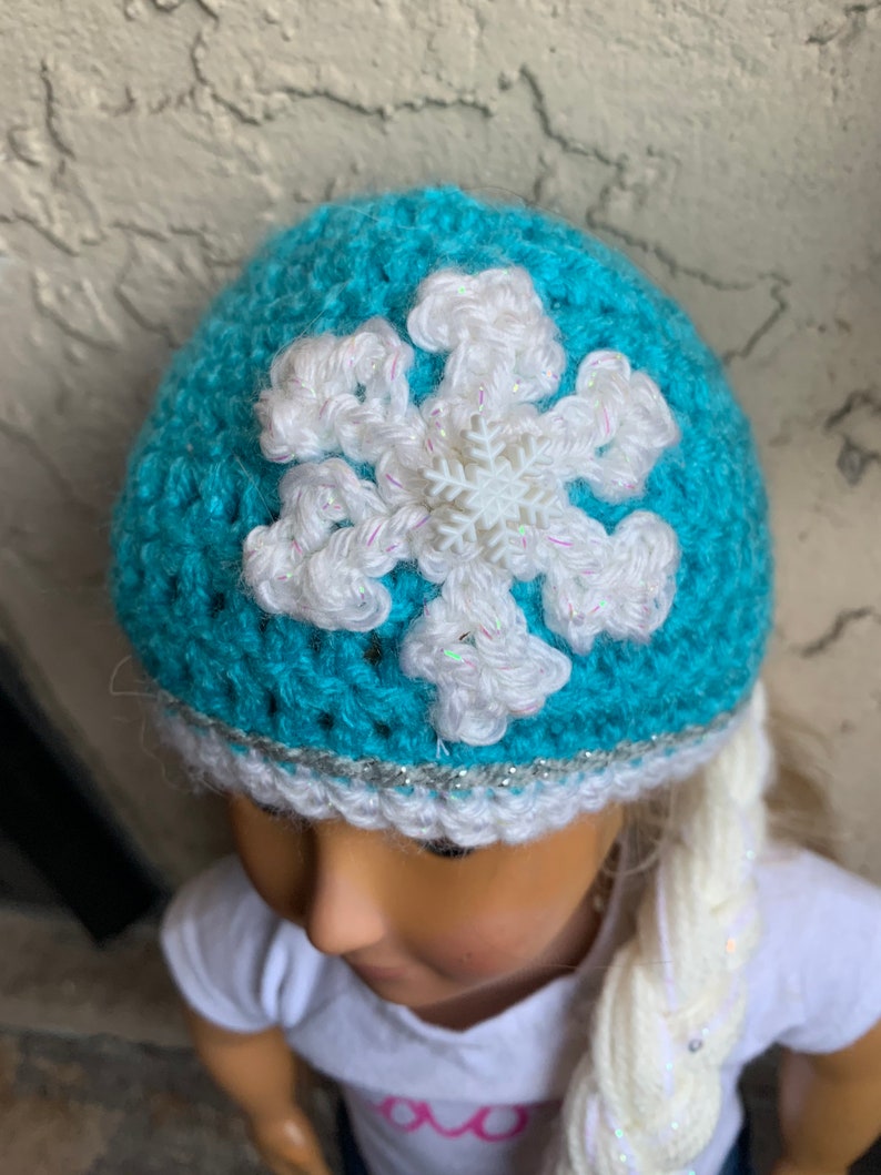 May include: A teal blue crocheted hat with a white snowflake design and a silver glitter trim. The hat is worn by a doll with long white braided hair.