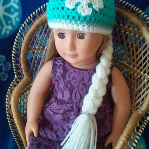 May include: A doll wearing a teal crocheted hat with a white snowflake design and a white crocheted braid.