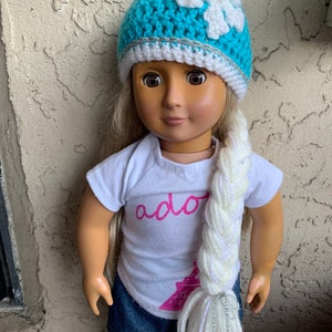 May include: A doll wearing a blue crocheted hat with a white snowflake design and a white and silver braided yarn tail. The doll is wearing a white t-shirt with the word "ador" printed on it and a blue denim skirt.