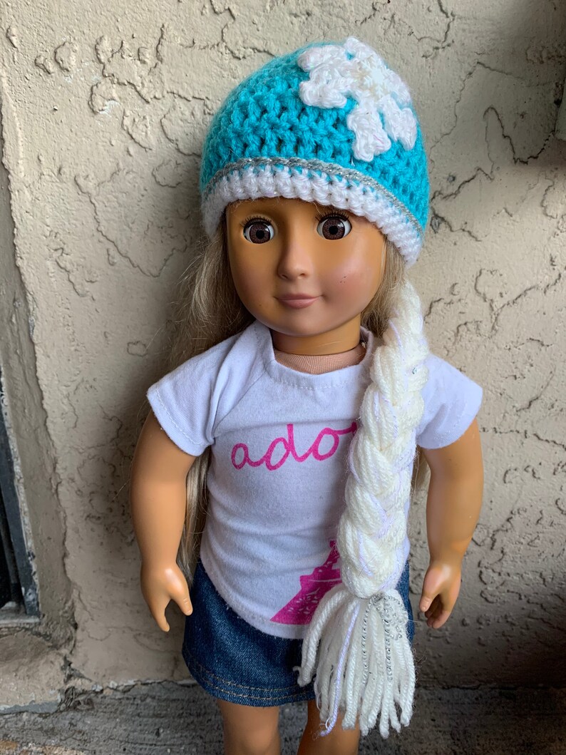 May include: A doll wearing a blue crocheted hat with a white snowflake design and a white and silver braided yarn tail. The doll is wearing a white t-shirt with the word "ador" printed on it and a blue denim skirt.