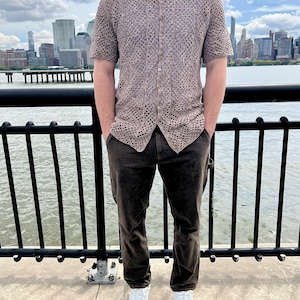 May include: A man wearing a brown open weave crochet shirt with a collared neckline, brown pants, and white sneakers. He is standing in front of a black metal railing with a city skyline in the background.
