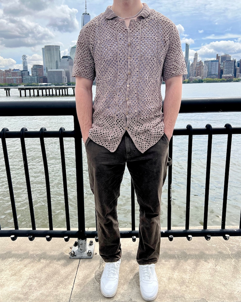 May include: A man wearing a brown open weave crochet shirt with a collared neckline, brown pants, and white sneakers. He is standing in front of a black metal railing with a city skyline in the background.