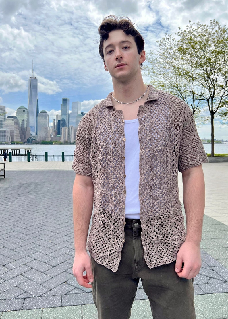 May include: A man wearing a brown crochet shirt with a white undershirt and green pants. He is standing in front of a city skyline.