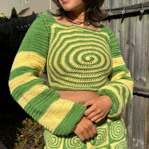 Puede incluir: Top de ganchillo verde y amarillo con un diseño en espiral en la parte delantera y las mangas. El top tiene un escote cuadrado y mangas largas con puños acampanados. La falda a juego es visible en la parte inferior de la imagen.