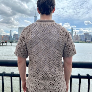 May include: A person wearing a brown crocheted shirt with a short sleeve design. The shirt has a loose fit and a square pattern. The person is standing on a pier overlooking a city skyline.