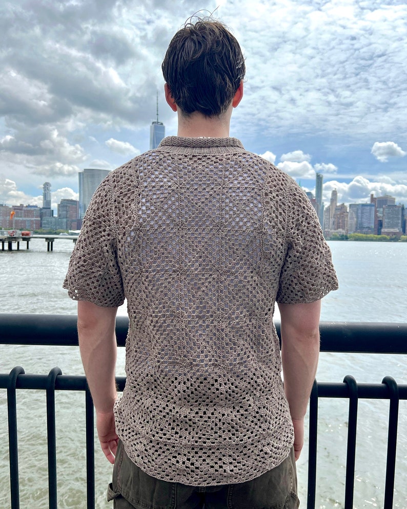 May include: A person wearing a brown crocheted shirt with a short sleeve design. The shirt has a loose fit and a square pattern. The person is standing on a pier overlooking a city skyline.