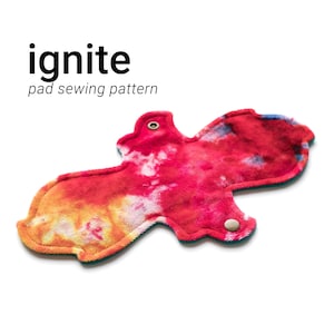 以下が含まれることがあります： 赤、オレンジ、青のタイダイ柄の布製パッドで、ターコイズブルーの縁取りと白いスナップボタンが2つ付いています。パッドは縫製用で、「ignite pad sewing pattern」とラベル付けされています。