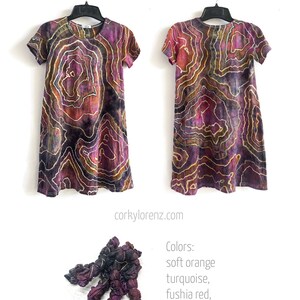 May include: A tie-dye dress with a swirling pattern in shades of purple, orange, and black. The dress has short sleeves and a round neckline. The dress is hanging on a hanger. The text "Colors: soft orange, turquoise, fushia red, raven." is below the dress.