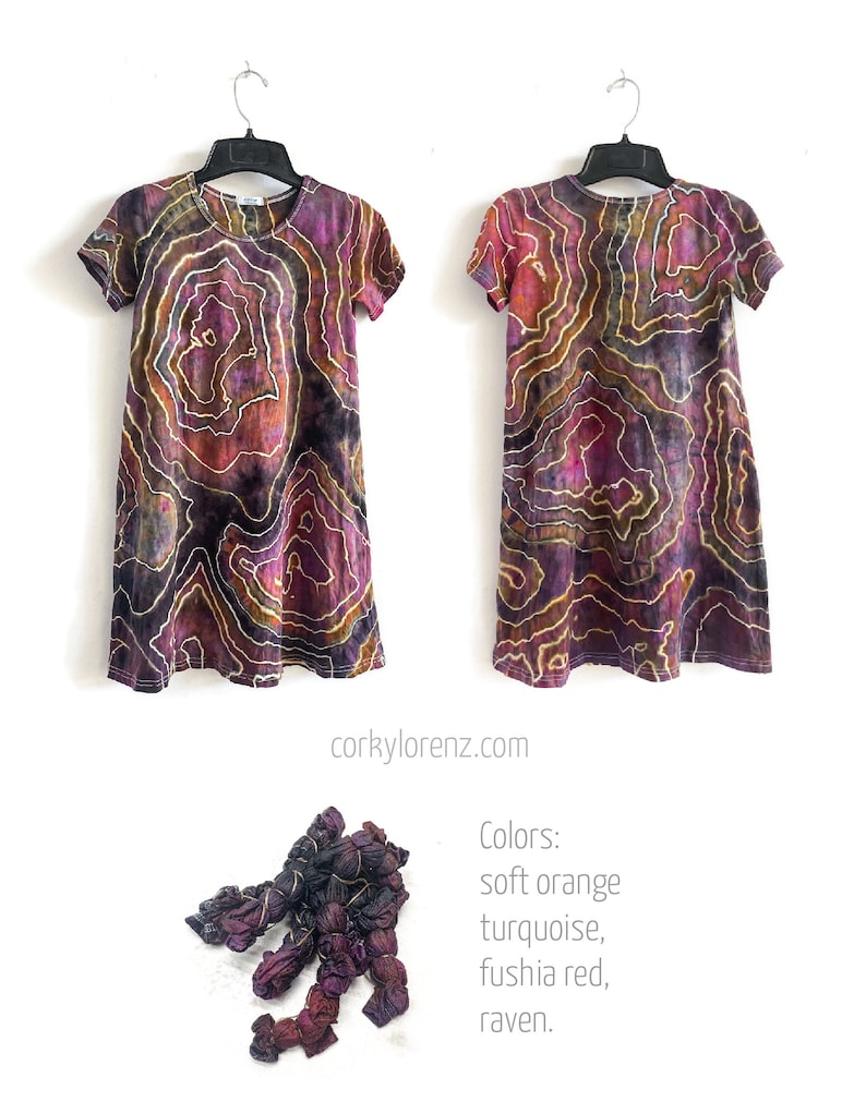May include: A tie-dye dress with a swirling pattern in shades of purple, orange, and black. The dress has short sleeves and a round neckline. The dress is hanging on a hanger. The text "Colors: soft orange, turquoise, fushia red, raven." is below the dress.