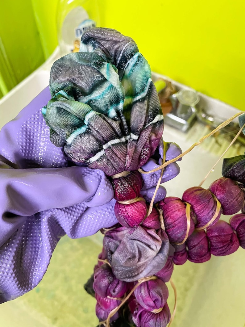 May include: A close-up of a purple fabric being tied with rubber bands. The fabric is being tied in a circular pattern, creating a series of small, round bundles. The fabric is a mix of purple, blue, and green colors.