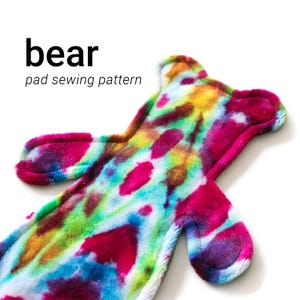 Può includere: Un modello di cucito a forma di orso con un design tie-dye. Il modello presenta tonalità vibranti di rosa, giallo, verde e blu. Il testo "bear pad sewing pattern" è visualizzato in nero nella parte superiore dell'immagine.