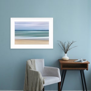 May include: A white framed photograph of a blurred seascape with blue and green water and a sandy beach. The photograph is hanging on a light blue wall above a white armchair with a beige throw blanket. A small wooden side table with a vase of dried branches sits to the right of the chair.