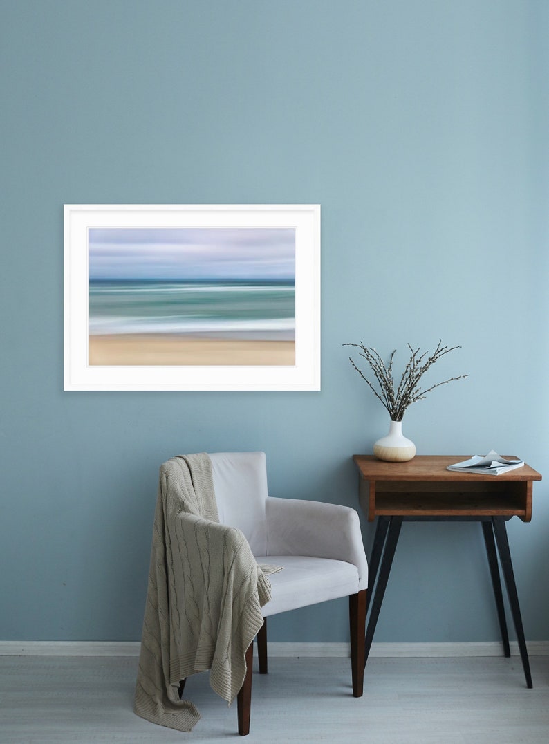 May include: A white framed photograph of a blurred seascape with blue and green water and a sandy beach. The photograph is hanging on a light blue wall above a white armchair with a beige throw blanket. A small wooden side table with a vase of dried branches sits to the right of the chair.