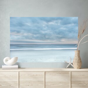 May include: A photograph of a beach scene with a cloudy sky and a calm sea. The sand is white and the water is a light blue. The photo is framed and hanging on a wall above a wooden dresser.