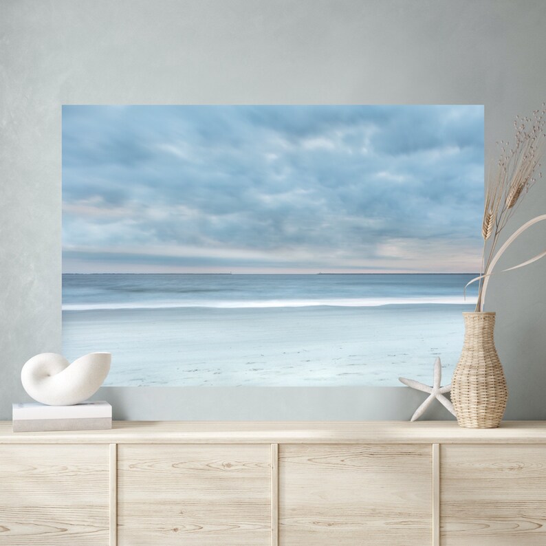 May include: A photograph of a beach scene with a cloudy sky and a calm sea. The sand is white and the water is a light blue. The photo is framed and hanging on a wall above a wooden dresser.