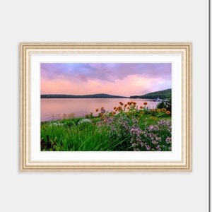 May include: A framed photograph of a lake with a mountain range in the background. The lake is calm and the sky is a soft pink and purple. There are flowers in the foreground.