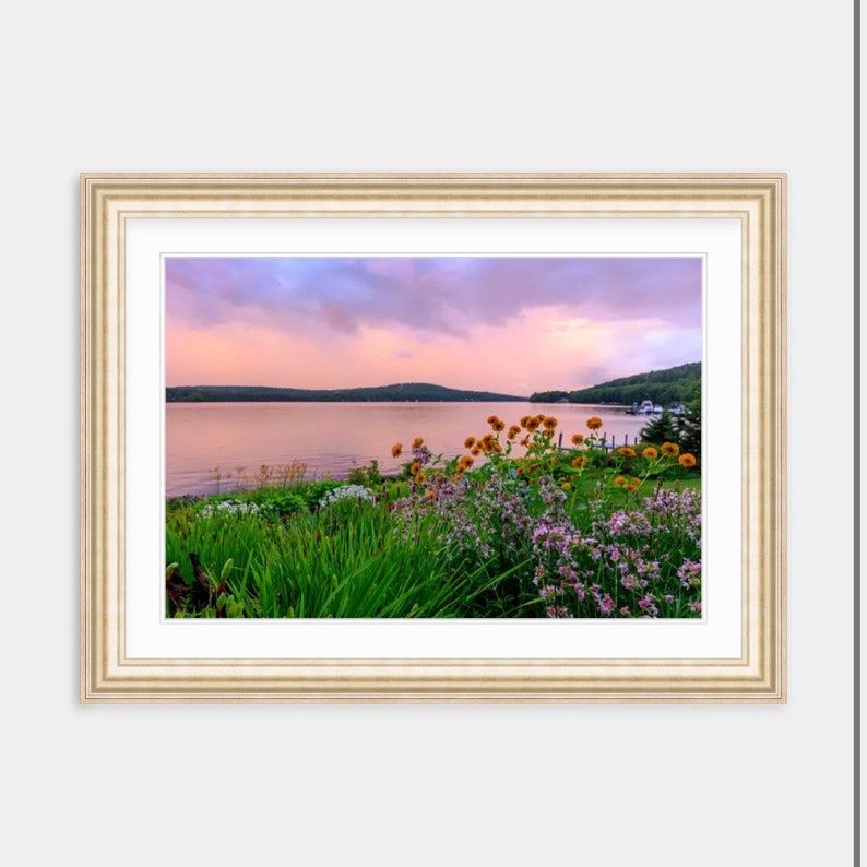 May include: A framed photograph of a lake with a mountain range in the background. The lake is calm and the sky is a soft pink and purple. There are flowers in the foreground.