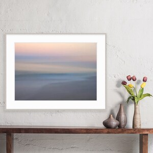 May include: A framed photograph of a blurred, abstract landscape with a soft, pastel color palette of pink, purple, and blue. The image depicts a beach scene with a horizon line and a sense of movement.