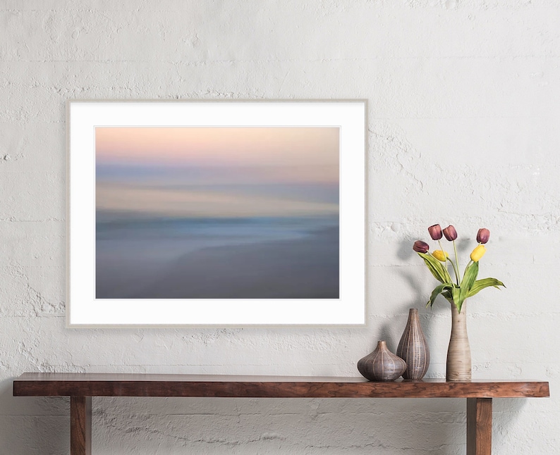 May include: A framed photograph of a blurred, abstract landscape with a soft, pastel color palette of pink, purple, and blue. The image depicts a beach scene with a horizon line and a sense of movement.