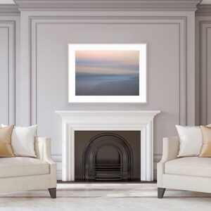 May include: Two white armchairs with gold throw pillows are positioned in front of a fireplace with a black fire grate. A framed photograph of a blurred seascape hangs above the fireplace.
