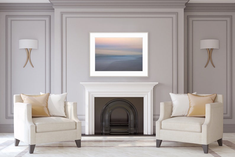 May include: Two white armchairs with gold throw pillows are positioned in front of a fireplace with a black fire grate. A framed photograph of a blurred seascape hangs above the fireplace.