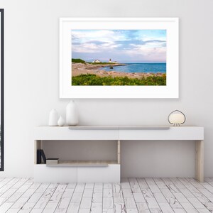 May include: A framed photograph of a lighthouse on a rocky coastline with a sandy beach and blue water in the background. The sky is a light blue with white clouds.