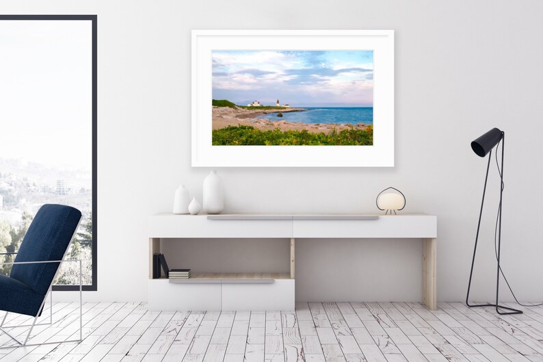 May include: A framed photograph of a lighthouse on a rocky coastline with a sandy beach and blue water in the background. The sky is a light blue with white clouds.