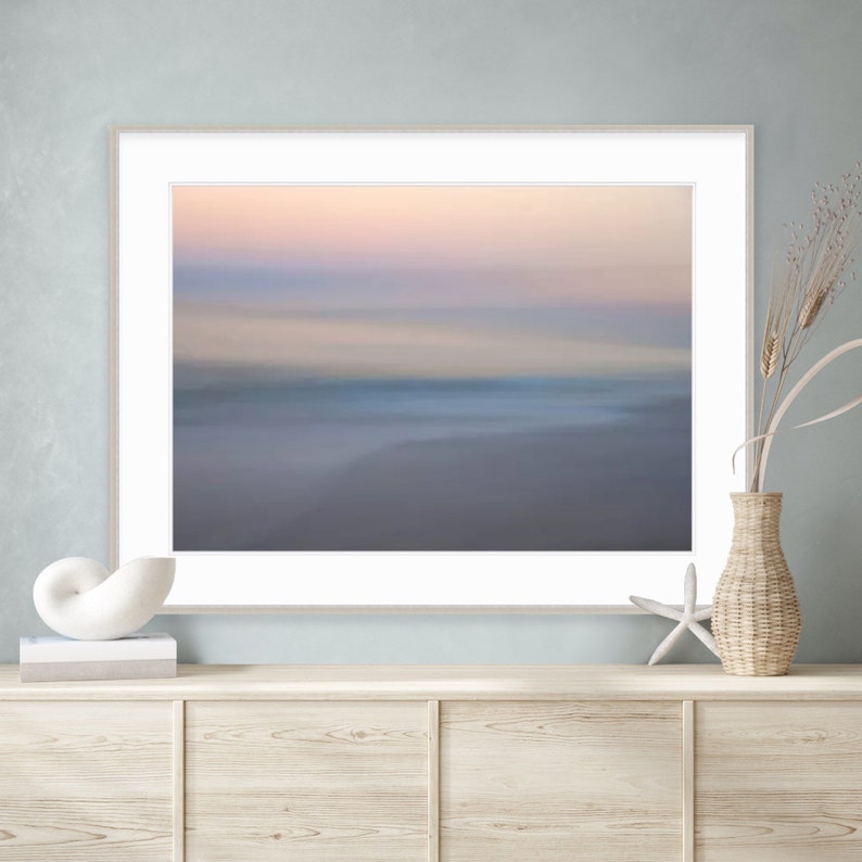 May include: A framed abstract photograph of a blurred beach scene at sunset. The colors are soft and muted, with shades of pink, purple, and blue. The image is framed in a simple white frame.
