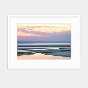 May include: A photograph of a beach at sunset with a winding creek in the foreground. The sky is a soft pink and purple, and the water is calm and reflective.