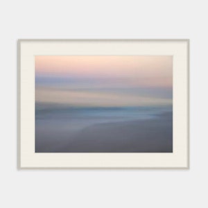 May include: A blurred abstract photograph of a beach scene at sunset. The sky is a soft pink and purple, and the water is a light blue. The sand is a light brown.