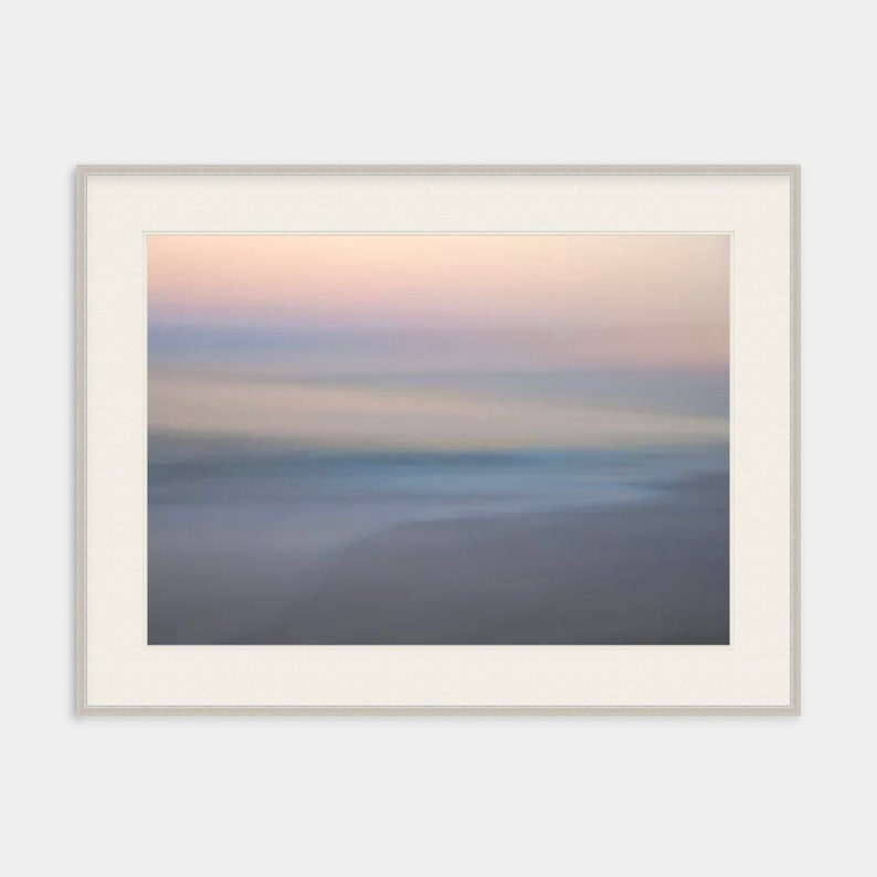 May include: A blurred abstract photograph of a beach scene at sunset. The sky is a soft pink and purple, and the water is a light blue. The sand is a light brown.