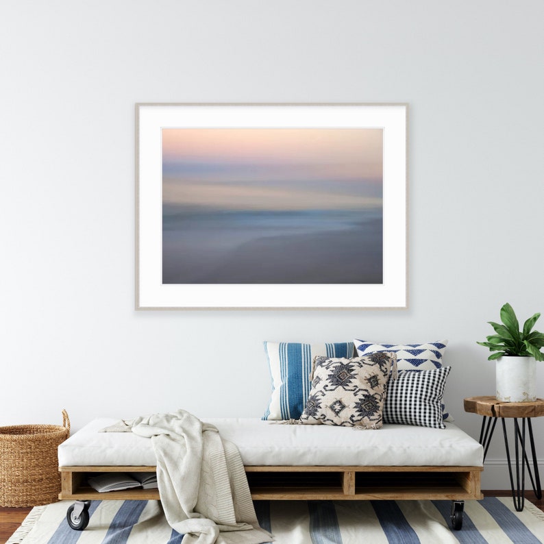 May include: A framed photograph of a blurred seascape with a soft pink and blue gradient sky. The image is displayed on a white wall above a white couch with blue and white patterned pillows.