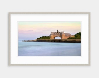 May include: A large, stone building with an arched entrance sits on a small island in the ocean. The sky is a soft pink and orange, and the water is a calm blue.