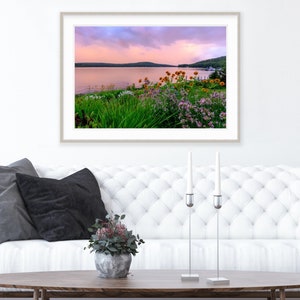 May include: A framed photograph of a lake with a pink and purple sunset sky. The lake is surrounded by green trees and bushes with yellow and purple flowers in the foreground.
