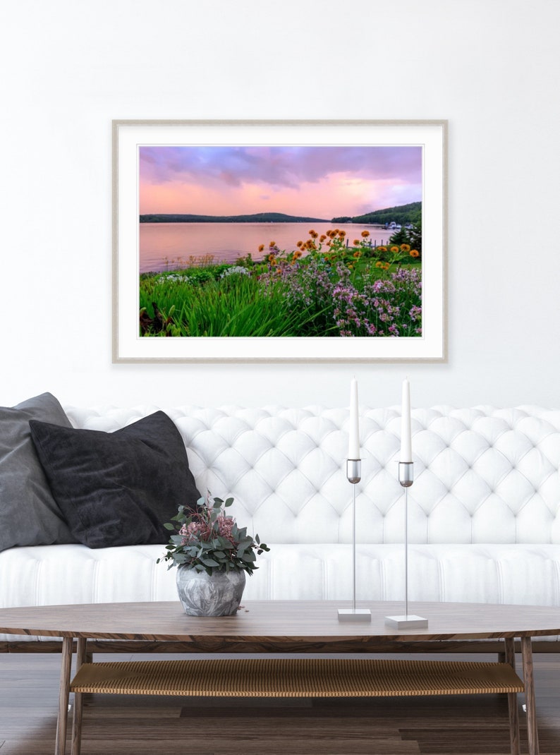 May include: A framed photograph of a lake with a pink and purple sunset sky. The lake is surrounded by green trees and bushes with yellow and purple flowers in the foreground.