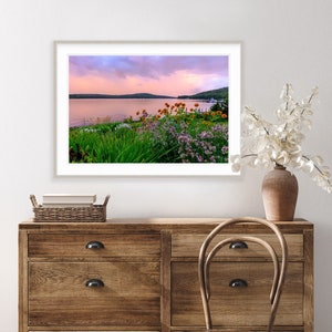 May include: A framed photograph of a lake with a pink and purple sunset sky. The lake is surrounded by green trees and bushes. There are wildflowers in the foreground.