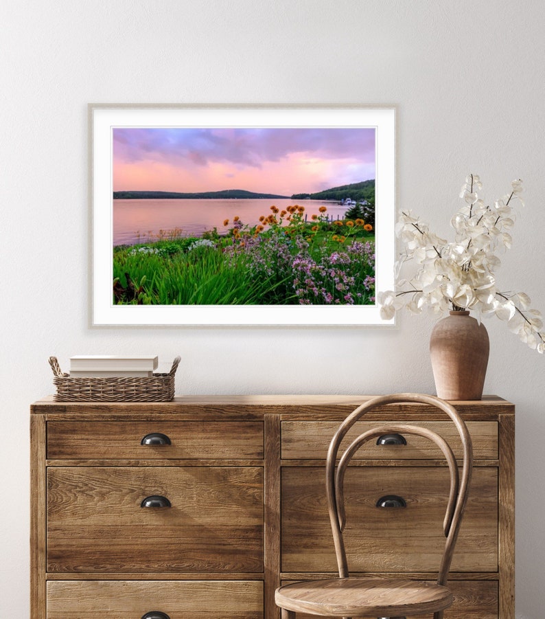 May include: A framed photograph of a lake with a pink and purple sunset sky. The lake is surrounded by green trees and bushes. There are wildflowers in the foreground.