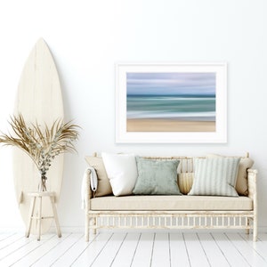 May include: A white couch with beige cushions and a white throw blanket sits in a room with white walls and wood floors. A surfboard leans against a white stool and a vase with dried branches sits on the stool. A framed print of a blurred beach scene hangs on the wall above the couch.
