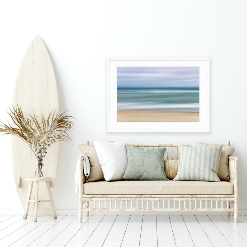 May include: A white couch with beige cushions and a white throw blanket sits in a room with white walls and wood floors. A surfboard leans against a white stool and a vase with dried branches sits on the stool. A framed print of a blurred beach scene hangs on the wall above the couch.
