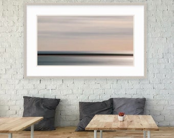 May include: A framed abstract landscape photograph with a blurred horizon and a light blue and gray color scheme. The photograph is hanging on a white brick wall above a wooden bench with gray cushions.