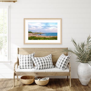 May include: A framed photograph of a lighthouse on a rocky coastline with a blue sky and white clouds. The lighthouse is white with a red roof and is located on a small island. The water is blue and the sky is a light blue with white clouds. The photo is framed in a light wood frame.