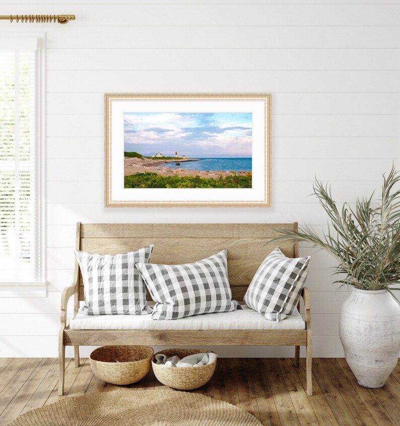 May include: A framed photograph of a lighthouse on a rocky coastline with a blue sky and white clouds. The lighthouse is white with a red roof and is located on a small island. The water is blue and the sky is a light blue with white clouds. The photo is framed in a light wood frame.