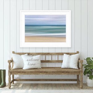 May include: A white framed print of a blurred seascape with blue and green water and a sandy beach. The print is hanging on a white shiplap wall above a wooden bench with two pillows and a green plant in a white pot.