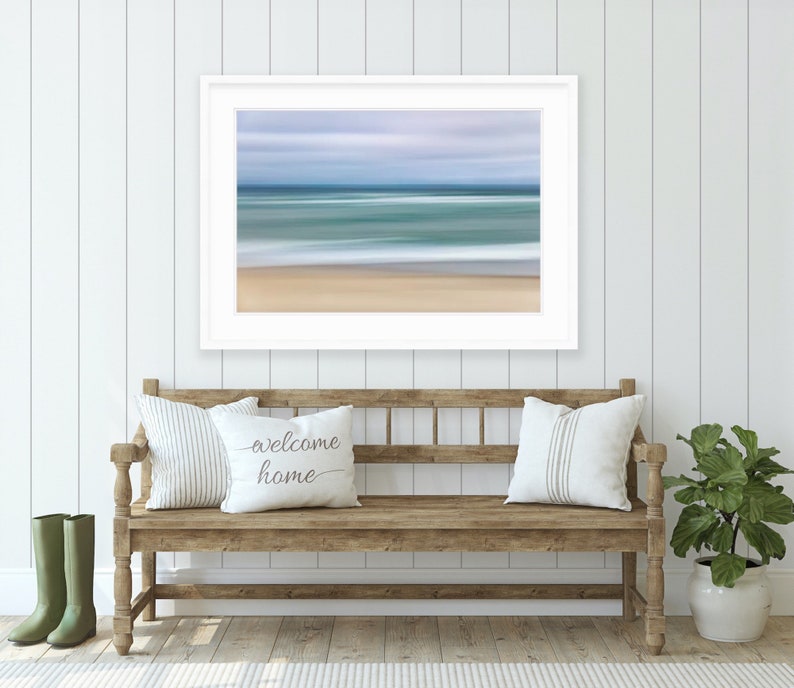 May include: A white framed print of a blurred seascape with blue and green water and a sandy beach. The print is hanging on a white shiplap wall above a wooden bench with two pillows and a green plant in a white pot.