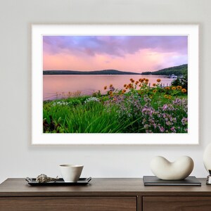 May include: A photograph of a lake with a pink and purple sunset sky. The lake is surrounded by green trees and bushes with yellow and purple flowers in the foreground.