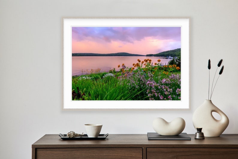 May include: A photograph of a lake with a pink and purple sunset sky. The lake is surrounded by green trees and bushes with yellow and purple flowers in the foreground.